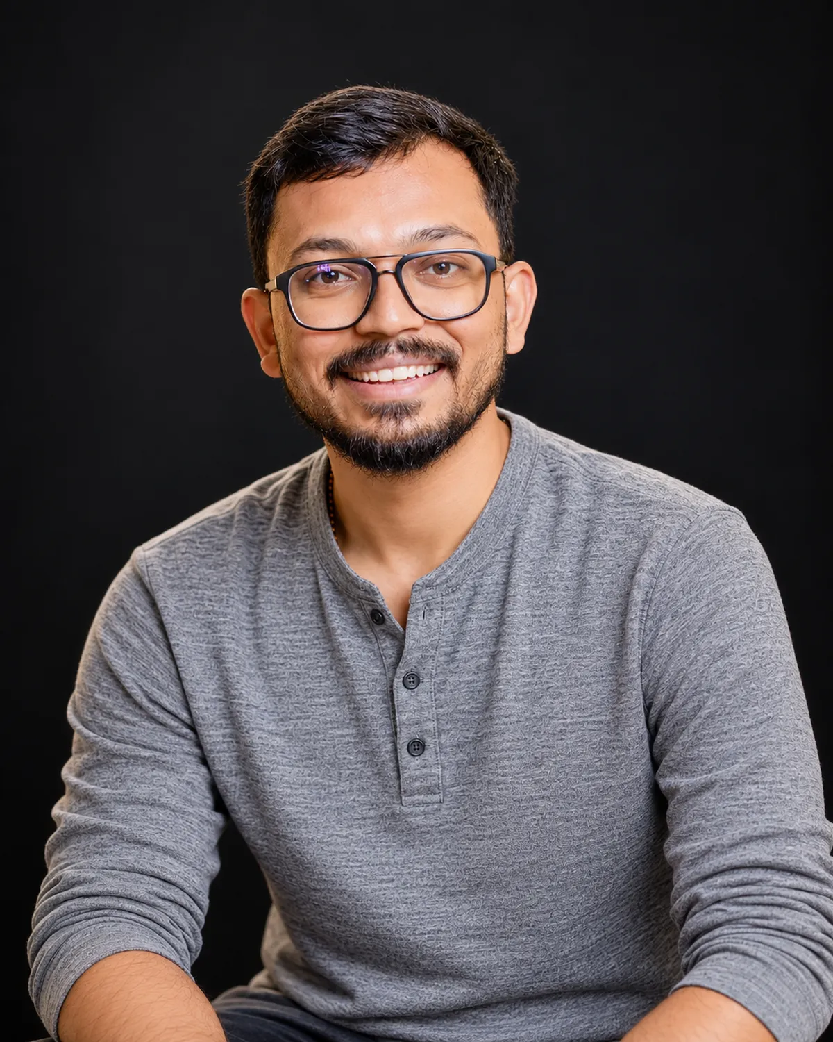 Tanishq Jain, smiling, in a grey henley against a black background.