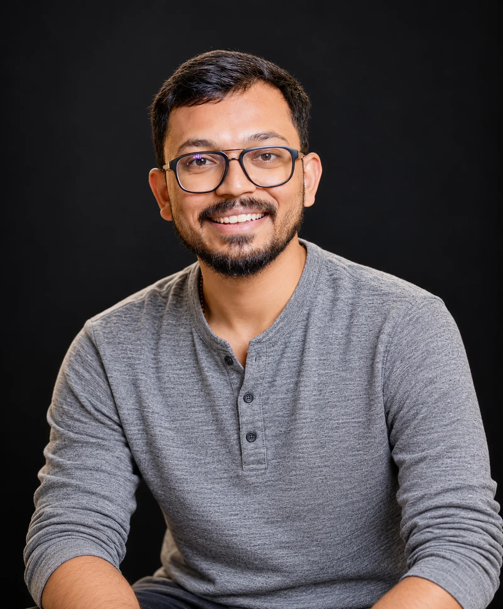 Tanishq Jain, smiling, in a grey henley against a black background.
